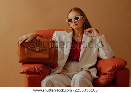 Fashionable confident woman wearing elegant white suit, sunglasses, chunky chain, holding classic brown leather bag, sitting in armchair, posing on beige background. Copy, empty space for text