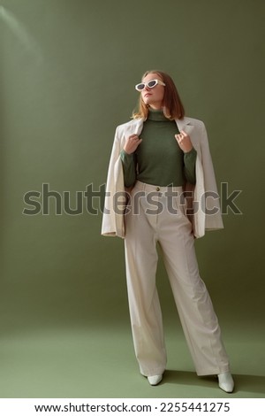 Fashionable confident woman wearing elegant white suit with blazer, wide leg trousers, cashmere turtleneck sweater, trendy sunglasses, posing on green background. Full-length studio fashion portrait