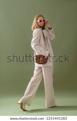 Fashionable confident woman wearing elegant white suit with blazer, wide leg trousers, trendy sunglasses, brown suede shoulder bag, posing on green background. Full-length studio fashion portrait