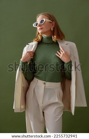 Fashionable confident woman wearing elegant white suit with blazer, trousers, cashmere turtleneck sweater, trendy cat eye sunglasses, posing on green background. Studio fashion portrait