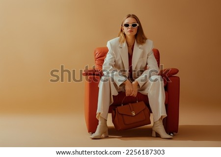 Fashionable confident woman wearing elegant white suit, sunglasses, ankle boots, holding classic brown leather bag, sitting in armchair, posing on beige background. Copy, empty space for text