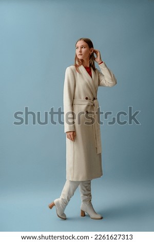 Fashionable confident woman wearing elegant white woolen coat,  high leather heeled boots, posing on blue background. Full-length studio fashion portrait. Copy, empty space for text
