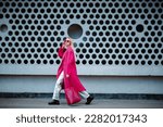 Fashionable confident woman wearing fuchsia color midi coat, sunglasses, metallic pants, combat boots, holding faux leather tote, shopper bag, walking in street of city. Copy, empty space for text