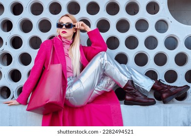 Fashionable confident woman wearing fuchsia color coat, blue sunglasses, pink turtleneck, metallic pants, combat boots, with  faux leather tote bag. Outdoor portrait. Copy, empty space for text