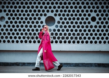 Fashionable confident woman wearing fuchsia color midi coat, sunglasses, metallic pants, combat boots, holding faux leather tote, shopper bag, walking in street of city. Copy, empty space for text