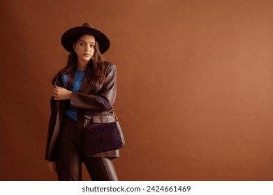 Fashionable confident woman wearing hat, faux leather suit blazer, pants, holding trendy purple purse, posing on brown background. Studio fashion portrait. Copy, empty, blank space for text - Powered by Shutterstock