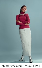 Fashionable confident woman wearing pink sunglasses, turtleneck, layered pearl necklace, white denim maxi skirt, silver boots, posing on blue background. Full-length studio fashion portrait