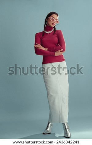 Fashionable confident woman wearing pink sunglasses, turtleneck, layered pearl necklace, white denim maxi skirt, silver boots, posing on blue background. Full-length studio fashion portrait