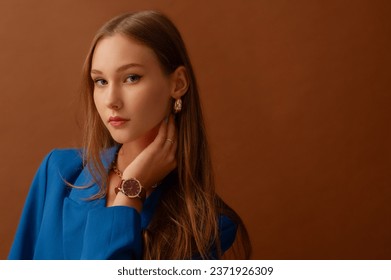 Fashionable confident woman wearing stylish dark red wrist watch, trendy blue suit blazer, golden hoop earrings. posing in studio, on brown backdrop. Copy, empty space for text