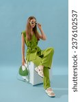 Fashionable confident woman wearing trendy green suit with waistcoat, trousers, white platform sandals, holding stylish quilted lather bag, posing in studio, on blue backdrop. Full-length portrait