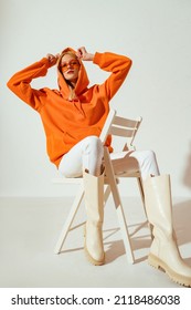 Fashionable confident woman wearing trendy orange hoodie, sunglasses, white skinny jeans, high boots, sitting, posing on chair. Full body portrait - Shutterstock ID 2118486038