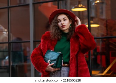 Fashionable confident woman wearing trendy winter orange faux fur coat, hat, green turtleneck sweater, holding stylish bag, posing in street of city.Copy, empty space for text