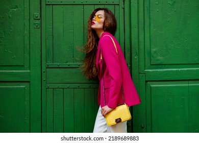 Fashionable confident woman wearing trendy outfit with yellow sunglasses, shoulder bag, pink fuchsia color blazer, posing near green door. Copy, empty space for text - Shutterstock ID 2189689883
