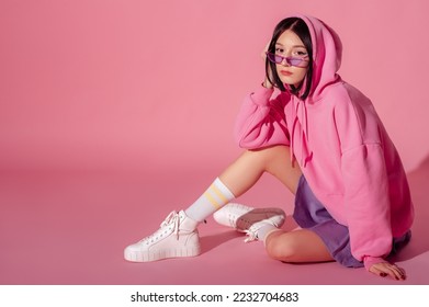 Fashionable confident woman wearing trendy pink hoodie, purple  sunglasses, mini skirt, white high top sneakers, striped socks, sitting, posing on pink background. Copy, empty space for text - Shutterstock ID 2232704683