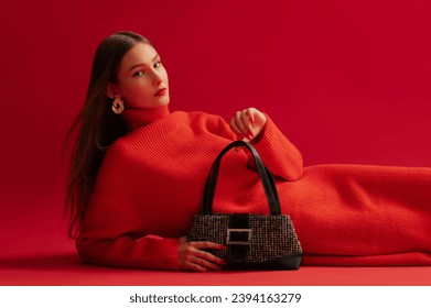 Fashionable confident woman wearing trendy red knitted turtleneck dress, golden earrings, holding baguette bag, posing, laying on red background. Studio fashion portrait. Copy, empty space for text - Shutterstock ID 2394163279