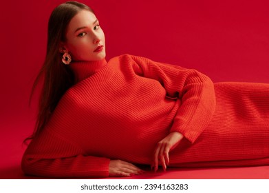 Fashionable confident woman wearing trendy red long knitted turtleneck dress, big golden earrings, posing, laying on red background. Studio fashion portrait. Copy, empty space for text - Shutterstock ID 2394163283