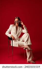 Fashionable confident woman wearing trendy white suit blazer, classic trousers, sunglasses, metallic color shoes, posing on red background