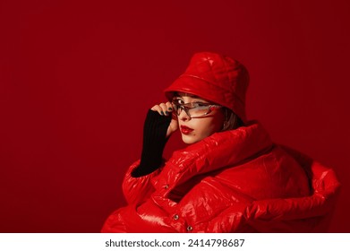 Fashionable confident woman wearing trendy y2k style silver glasses, quilted faux leather bucket hat, puffer jacket, posing on red background. Close up studio fashion portrait. Copy, empty space - Powered by Shutterstock