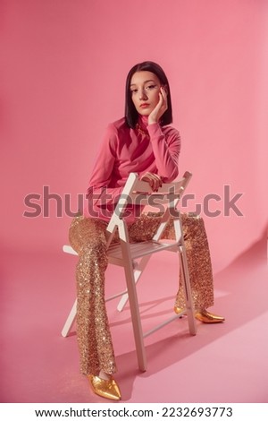 Fashionable confident woman wearing trendy 70s style outfit with  pink turtleneck top, sequined flare trousers, golden pointed toe shoes. Full-length studio portrait