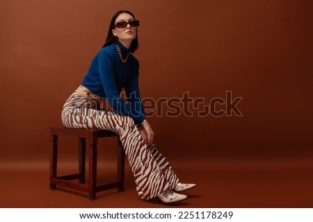 Fashionable confident woman wearing trendy brown rectangular sunglasses, stylish blue turtleneck, flared trousers with zebra print, boots. Full-length studio portrait. Copy, empty space for text