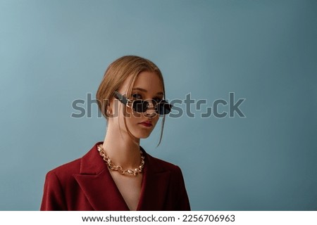 Fashionable confident woman wearing trendy black rectangular sunglasses, golden earrings, chunky chain, posing on blue background. Close up studio portrait. Copy, empty space for text