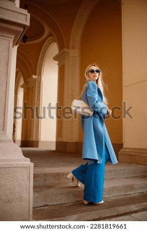 Fashionable confident woman wearing trendy outfit with blue coat, wide leg trousers, sunglasses, white ankle boots, quilted leather shoulder bag, posing in street. Full-length outdoor portrait