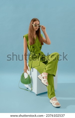 Fashionable confident woman wearing trendy green suit with waistcoat, trousers, white platform sandals, holding stylish quilted lather bag, posing in studio, on blue backdrop. Full-length portrait