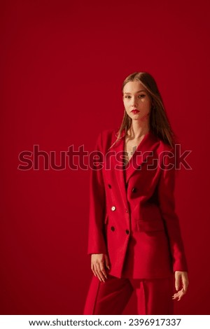 Fashionable confident woman wearing trendy red suit double breasted blazer, classic trousers, posing on red background. Studio fashion portrait. Copy, empty space for text