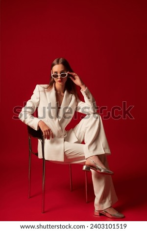 Fashionable confident woman wearing trendy white suit blazer, classic trousers, sunglasses, metallic color shoes, posing on red background