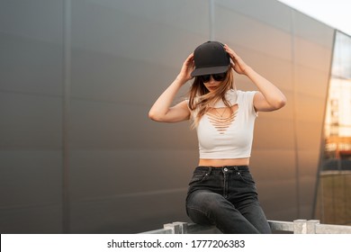 Fashionable cool hipster woman in sunglasses in black baseball cap in torn white t-shirt in vintage jeans with tattoo posing on dark background at sunset. Urban American girl rests in city. - Powered by Shutterstock