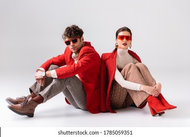 fashionable couple in red blazers and sunglasses sitting on floor while posing on grey
