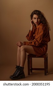 Fashionable Curly Brunette Woman Wearing Brown Cotton Mini Shirt Dress, Trendy Track Sole Leather Boots, Posing In Studio. Full-length Studio Portrait