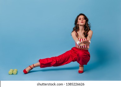 Fashionable Curly Woman From 80s Doing Stretching On Blue Background. Brunette In Red Pants And Striped Top Doing Fitness