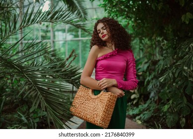 Fashionable curly woman wearing pink asymmetric ruffled blouse, holding straw wicker top handle bag, posing in tropical garden. Summer fashion, lifestyle conception. Copy, empty space for text  - Shutterstock ID 2176840399