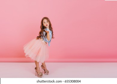 Fashionable cute young girl with long brunette hair, in stylish tulle skirt wearing big mother`s shoes, sending kiss to camera isolated on pink background. Having fun of joyful kid. Place for text