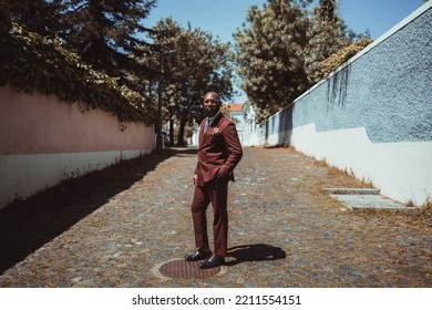 A Fashionable Elegant Bald Black Man Entrepreneur With A Well-groomed Beard, In A Tailored Suit Of A Chestnut Color, Is Standing On The Paving Stone In The Middle Of A Narrow Street On Warm Sunny Day