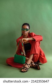 Fashionable elegant Black woman wearing trendy green sunglasses, orange suit with blazer, trousers, strap sandals, leather bag. Full-length studio portrait with natural day light