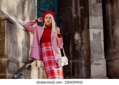 Fashionable elegant blonde woman wearing stylish red beret, turtleneck, classic pink suit blazer, checkered skirt, holding trendy bag, posing in street of European city. Copy, empty space for text - Shutterstock ID 2204359925