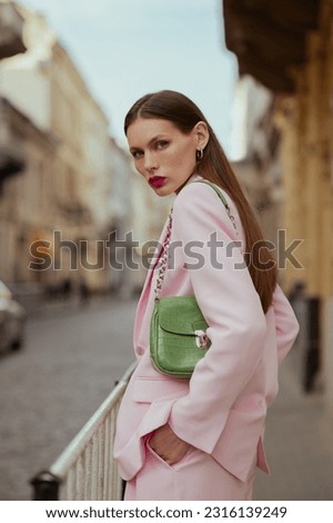 Fashionable elegant confident woman wearing trendy pink suit blazer, with green faux leather shoulder bag, walking in street of European city. Outdoor fashion portrait

