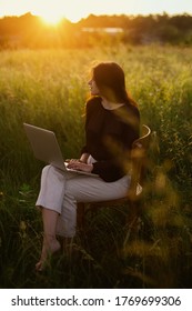 Fashionable Elegant Girl Working On Laptop And Sitting On Rustic Chair  In Sunny Summer Field At Sunset. New Office Concept.  Remote Work With Social Distancing And Safety Protocols