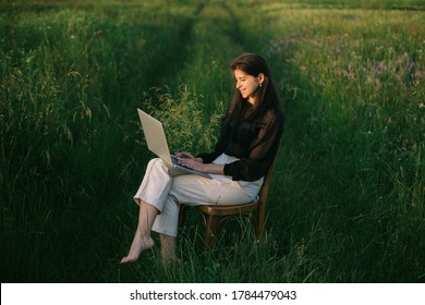 Fashionable Elegant Girl Working On Laptop And Sitting On Rustic Chair  In Green  Summer Field. New Office Concept. Remote Work With Social Distancing And Safety Protocols