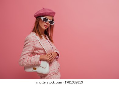 Fashionable, elegant woman wearing classic beret, tweed blazer, stylish rectangle white frame sunglasses, with white leather bag, posing on pink background. Copy, empty space for text 