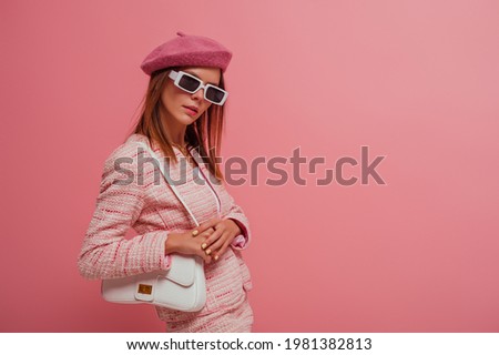 Fashionable, elegant woman wearing classic beret, tweed blazer, stylish rectangle white frame sunglasses, with white leather bag, posing on pink background. Copy, empty space for text 