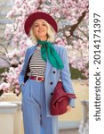 Fashionable, elegant woman wearing trendy blue suit, green neckscarf, burgundy color hat, holding big wicker leather bag, posing in street near blooming tree. Spring fashion, lifestyle conception