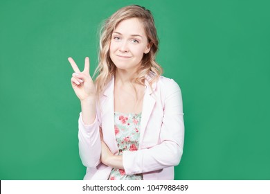 Fashionable female model with wavy hair, wears trendy colorful dress, round lips, gestures against green background, feels like star. Beautiful blonde woman shows peace sign. Body language concept. - Powered by Shutterstock