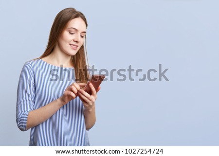 Fashionable female in stylish clothes, holds smart phone, checks her bank account, poses against blue background with copy space for your promotional text or advertisment. Technology and people