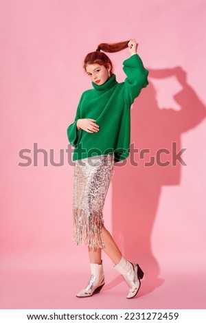 Fashionable freckled girl wearing trendy green knitted turtleneck sweater with wide sleeves, sequin skirt with fringes, stylish cowboy ankle boots. Full-length studio portrait
