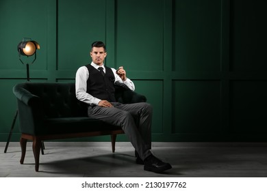 Fashionable gentleman sitting on sofa and holding watch in dark room