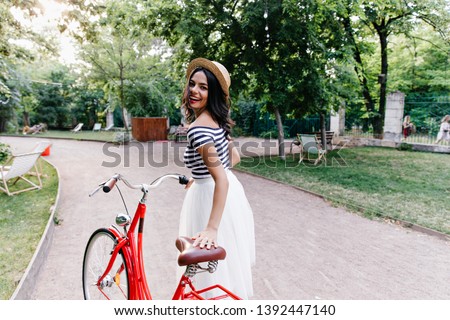 Fashionable girl in long white skirt having fun in summer weekend. Outdoor portrait of amazing brunette lady with bicycle looking over shoulder.