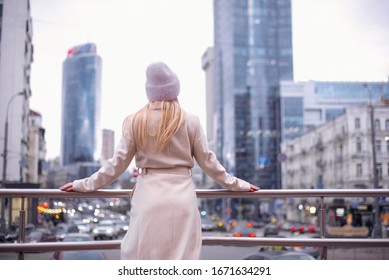 Fashionable Girl Looks At A Skyscraper, Rear View. Girl Wearing A Coat And A Pink Hat.